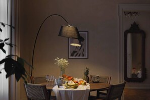 Un comedor con una mesa dispuesta para dos personas, un jarrón con flores, un frutero, una lámpara y un cuadro en la pared.