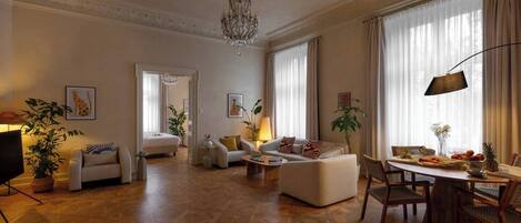 A spacious living room with a chandelier, a dining area with a table set for two, and a separate bedroom with a bed and a large window.