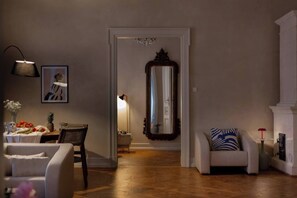 A living room with a wooden floor, a white armchair, a dining table with fruits, a framed picture, and a mirror.