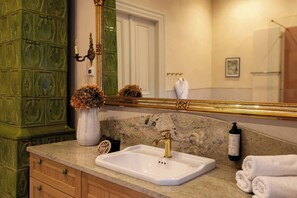Un baño con lavamanos de mármol, grifo dorado, espejo grande, mueble verde y un florero con flores secas.