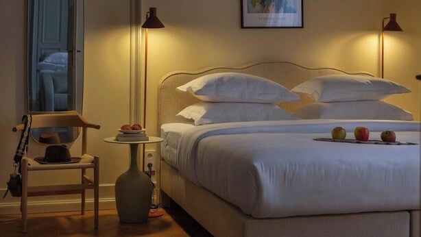 A neatly made bed with white linens, a small round table with books and a vase, and two floor lamps.