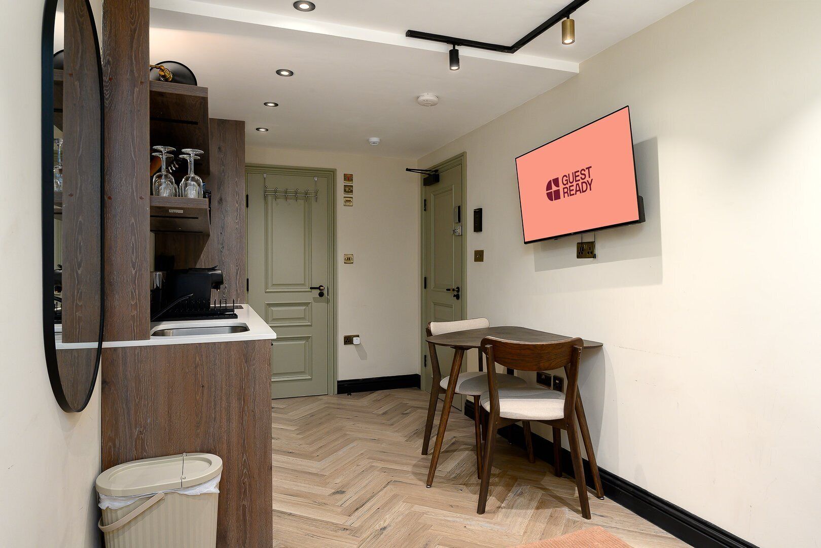 Ein Hotelzimmer mit einem hölzernen Schminktisch, einem Schreibtisch mit Stuhl, einem Fernseher der 'GUEST READY' anzeigt und einer Tür, die in einen anderen Raum führt.