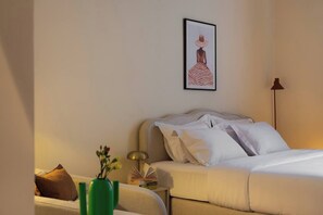 A neatly made bed with white linens, a framed artwork on the wall, a bedside lamp, and a green vase with flowers.