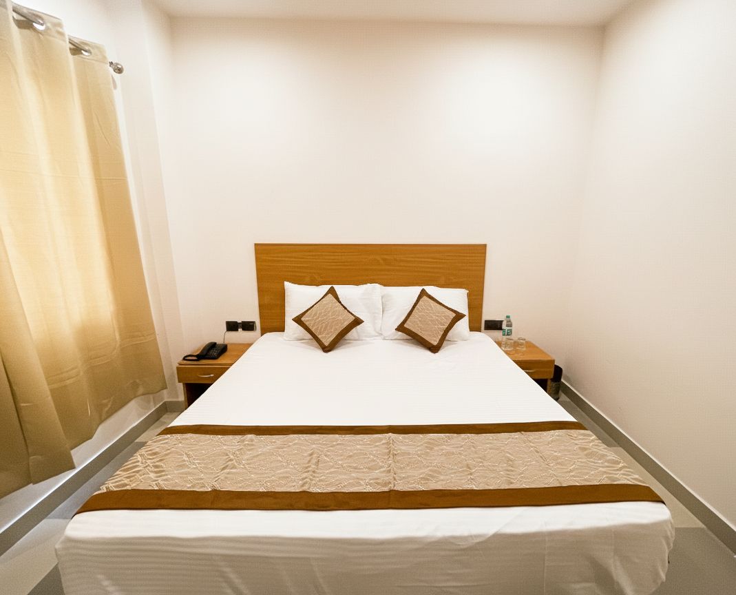 Una habitación de hotel con una cama, un cabecero de madera, dos almohadas, una mesita de noche con un teléfono y una mesita auxiliar con una botella.