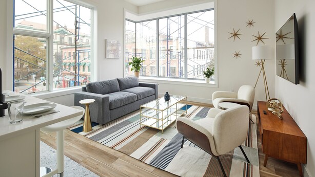 A modern living room with a sofa, coffee table, and chairs, large windows with city views, and a wooden sideboard. - Prime Hoboken: 2BR Loft by PATH (Hoboken)