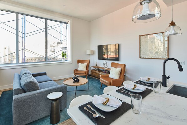 A modern living room with a grey sofa, two armchairs, a dining table set for two, and a large window overlooking a construction site.