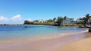 Una playa con agua cristalina, orilla de arena y edificios con palmeras al fondo.