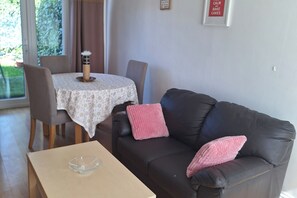 Un salón con un sofá marrón oscuro, una mesa redonda con mantel floreado y un cuenco de vidrio sobre una caja de cartón.