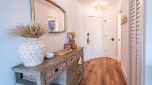 A hallway with a wooden floor, a mirror on the wall, a vase with dried plants, and a console table with baskets.