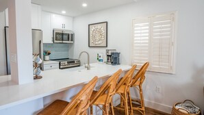 A modern kitchen with white cabinets, a stainless steel refrigerator, a microwave, and a coffee maker. There are wicker chairs around a white island.