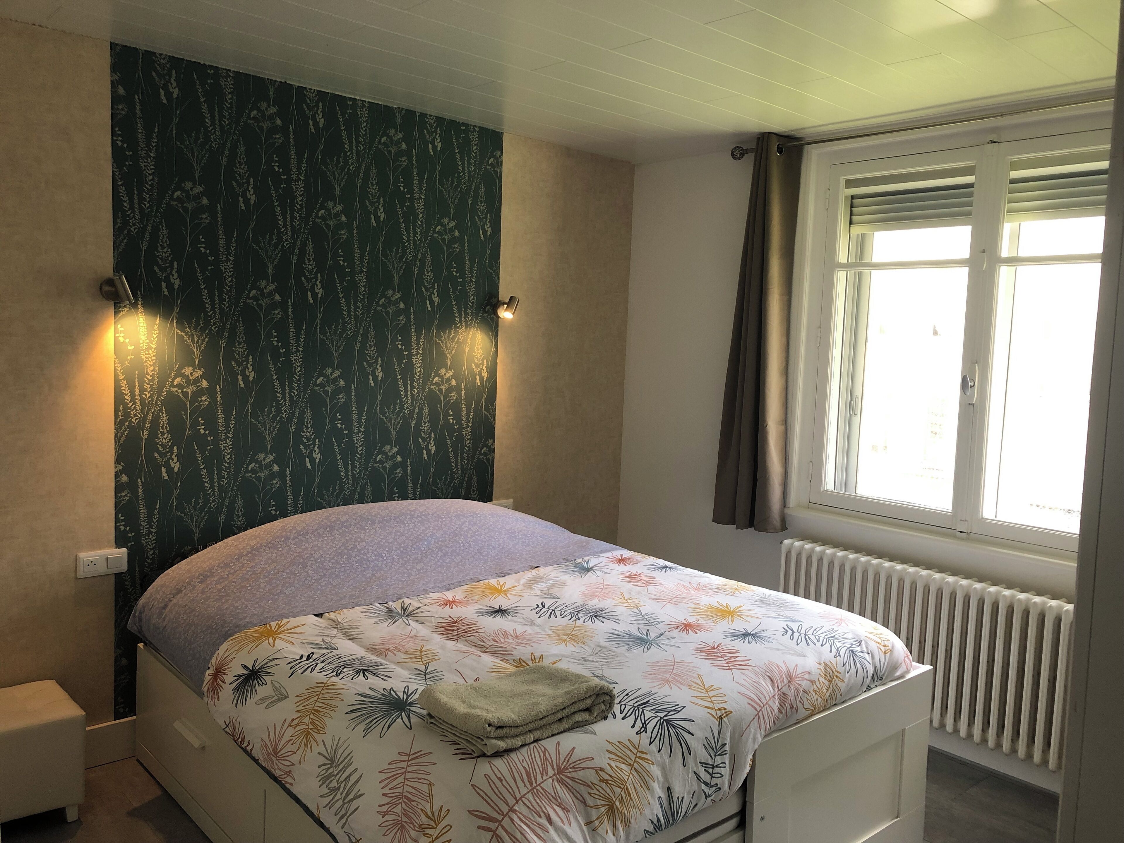 Een slaapkamer met een bed, een radiator, een raam met gordijnen en een bloemenbehang wand.