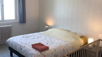 Ein Schlafzimmer mit einem Bett, einem Holckrippenbett, einer Pendelleuchte, einem Fenster mit Vorhängen und einer Heizung.
