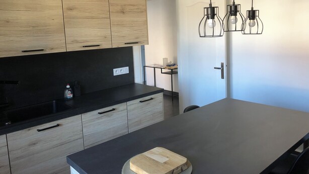 Una cocina moderna con armarios de madera, encimera negra y una isla central. Hay luces colgantes sobre la isla y una pequeña mesa con una botella de detergente.