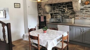 A dining area with a table set for four, a stone wall, and a kitchen with a sink and stove. - Cottage in the heart of the village of Kerascoët. (NEVEZ)