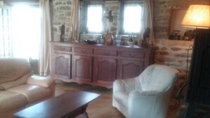 A living room with a sofa, armchair, and a coffee table. There is a stone wall and a fireplace with a lamp. - Cottage in the heart of the village of Kerascoët. (NEVEZ)