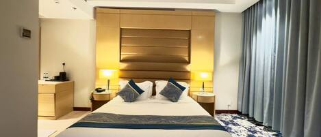 Una habitación de hotel con una cama grande, dos mesitas de noche, un cabecero y una ventana con cortinas.