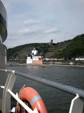 Blick auf ein historisches Gebäude mit einem Turm und einer Burg auf einem Hügel, aufgenommen vom Wasser aus.
