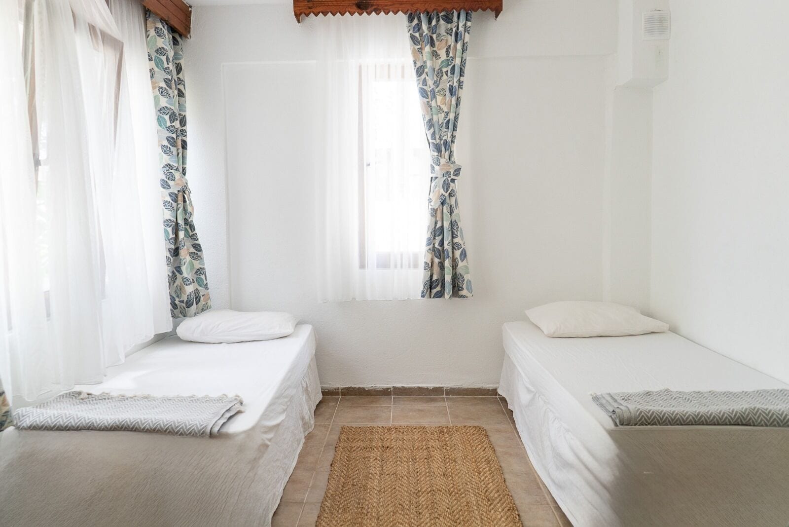 A small, clean room with two single beds, a window with sheer curtains, and a woven rug on the floor.