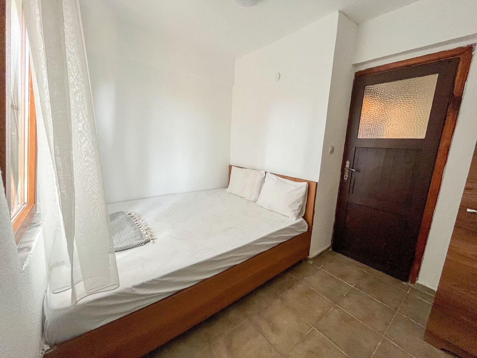A small, single-bed room with a wooden headboard, a plain white wall, and a door with a textured glass panel.