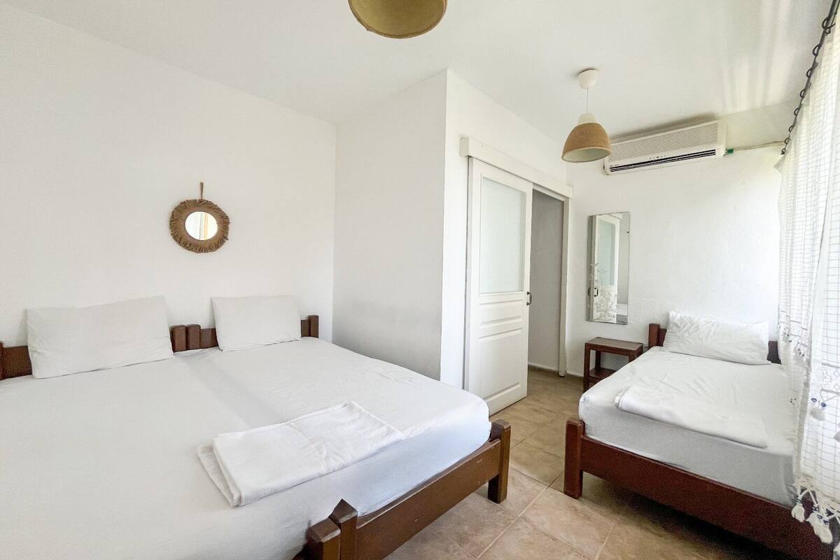 Quarto de hotel com duas camas, um espelho redondo, um ventilador de teto e uma porta que dá para outro quarto.