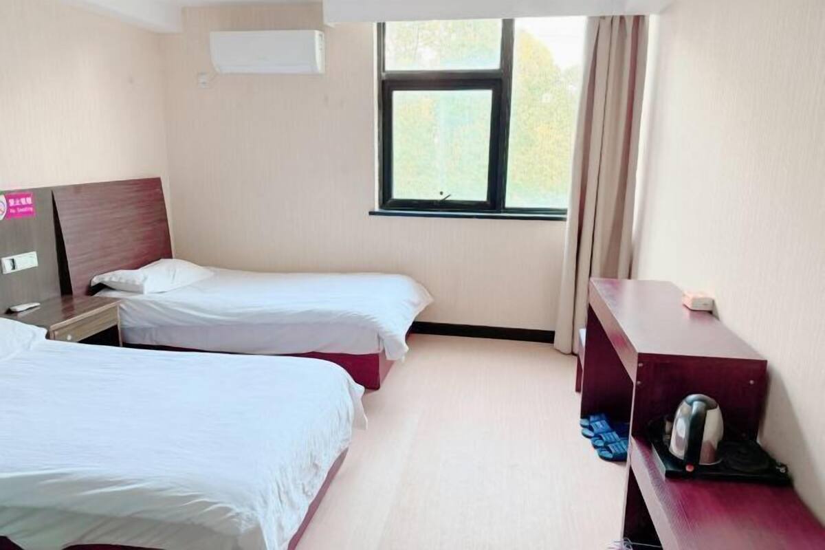 A hotel room with two beds, a desk, a window with curtains, and a kettle on the desk.