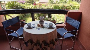 A round table with a floral tablecloth, two blue chairs, a pitcher, and two cups on a balcony with a view of a garden. - Villetta Kala e Moru "Sa Oghe e su Mare" (Quartu Sant'Elena)