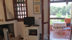A room with a television on a white cabinet, a window with a wooden frame, and a view of greenery outside. - Villetta Kala e Moru "Sa Oghe e su Mare" (Quartu Sant'Elena)