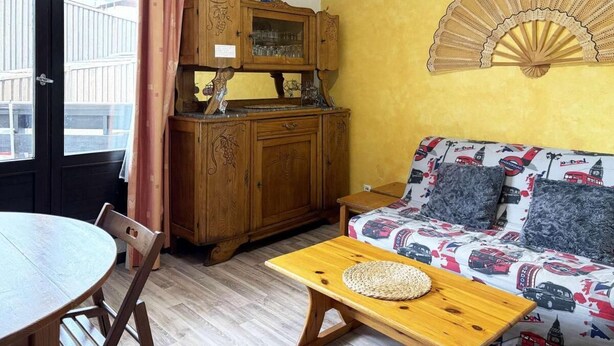 Una habitación con un armario de madera, un sofá estampado con motivos de autobús, un ventilador, una mesa redonda y una ventana con cortina.