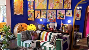 Ein lebendiges Wohnzimmer mit blauer Wand, farbenfroher abstrakter Kunst, einem gestreiften Sofa, einem Couchtisch mit Büchern und einem großen Spiegel.