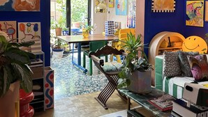Ein lebendiger Raum mit blauer Wand, farbenfroher Kunst, einem Essbereich und einer gemütlichen Sitzecke mit Büchern und Pflanzen.