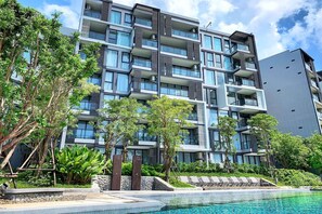 A modern multi-story building with balconies, a swimming pool, and lush greenery.