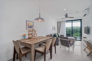 A modern living room with a dining area, a ceiling fan, and a large window with a view of greenery.