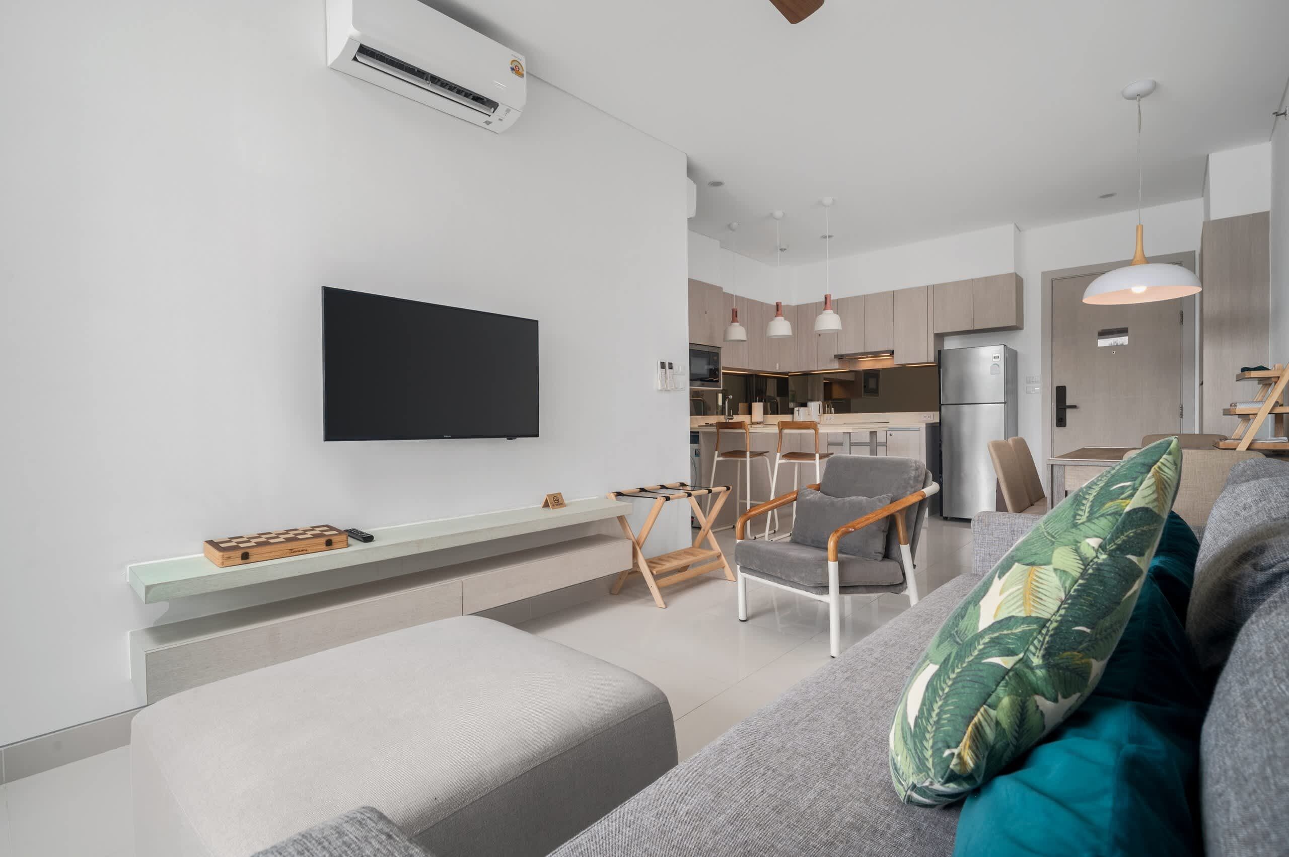 A modern living room with a flat-screen TV, a sofa, a coffee table, and a dining area with chairs and a kitchen in the background.
