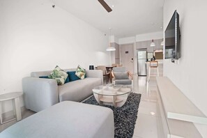 Una sala moderna con sofá, reposapiés y mesa de centro, un área de cocina al fondo y un televisor de pantalla plana montado en la pared.