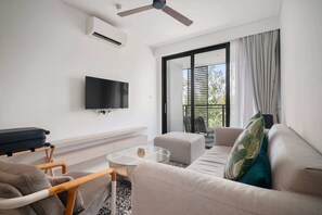 A modern living room with a flat-screen TV, a ceiling fan, a sofa, a glass coffee table, and a view of the outdoors through a sliding glass door.