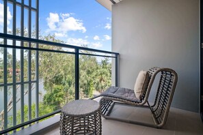 Un balcon avec une chaise en osier et un tabouret rond, donnant sur une étendue d’eau et une zone verdoyante.