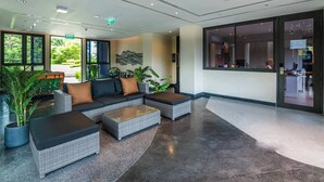 A lobby with a sectional sofa, two side tables, potted plants, and a large window offering a view of the outdoors.