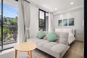 Quarto moderno com sofá-cama, mesa de madeira redonda e varanda com vista para o verde.