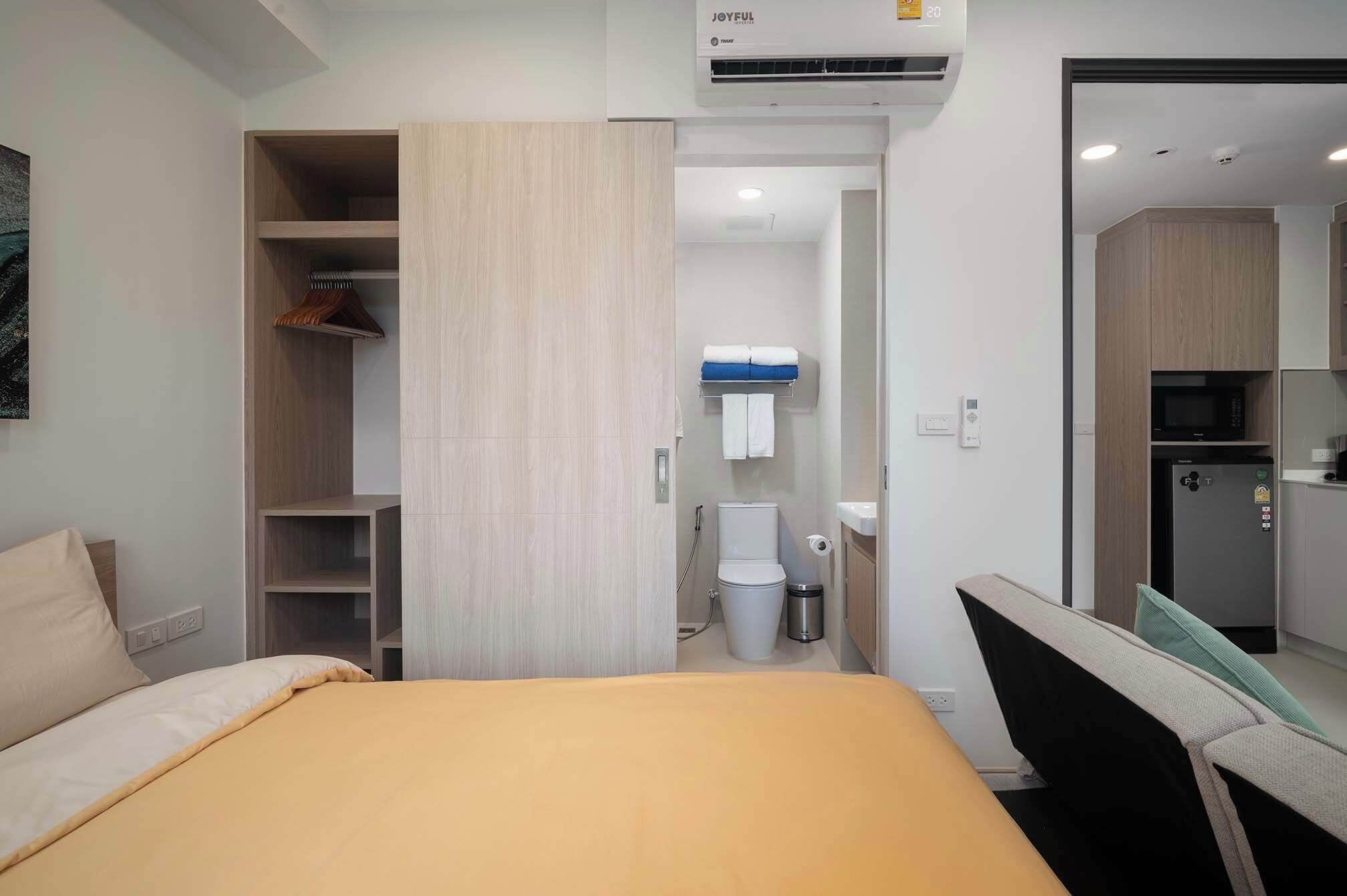 Quarto de hotel com cama, guarda-roupa, banheiro com vaso sanitário e cozinha compacta.