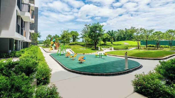 A playground with swings and a slide, surrounded by greenery and a building.