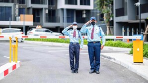 Deux personnes en uniforme se tiennent devant une entrée fermée par un portail, avec un bâtiment moderne en arrière-plan.