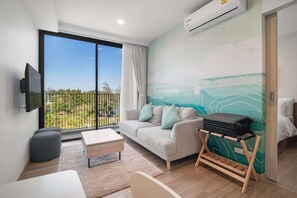 Ein modernes Hotelzimmer mit großem Balkon, einer Couch mit Kissen, einem kleinen Tisch, einem Bett und einem an der Wand befestigten Fernseher.