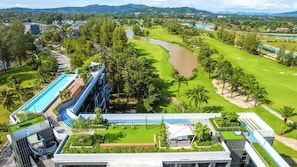 A golf course with a water hazard, a swimming pool, and a modern building with a rooftop garden. - Skypark Aurora 2, Two-Bedroom 2Cx1 Premium (Choeng Thale)