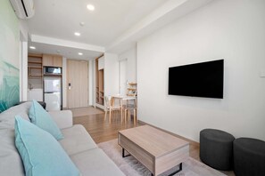 A modern living room with a sofa, coffee table, and ottomans. There is a TV mounted on the wall and a kitchen area visible in the background.