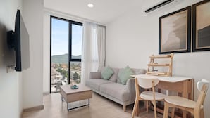 Un salon moderne comprenant un canapé, une table basse, une table à manger avec des chaises, un téléviseur fixé au mur et de grandes fenêtres offrant une vue sur les bâtiments et les collines.