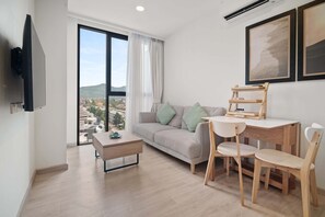A modern living room with a sofa, a coffee table, a dining table with chairs, a TV mounted on the wall, and large windows with a view of buildings and hills. - Skypark Aurora, One Bedroom 3709 1Bx Premium (Choeng Thale)
