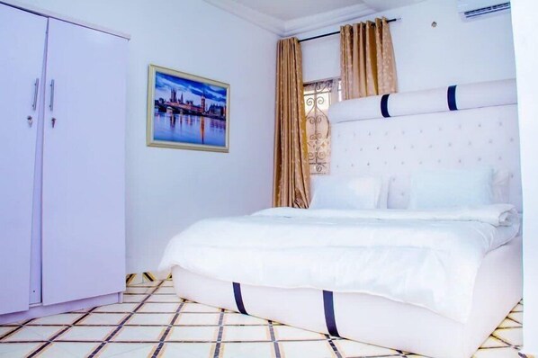 A bedroom with a bed, a wardrobe, a framed picture on the wall, and a window with curtains. - Hollysee Hotel and Suites (Lagos)