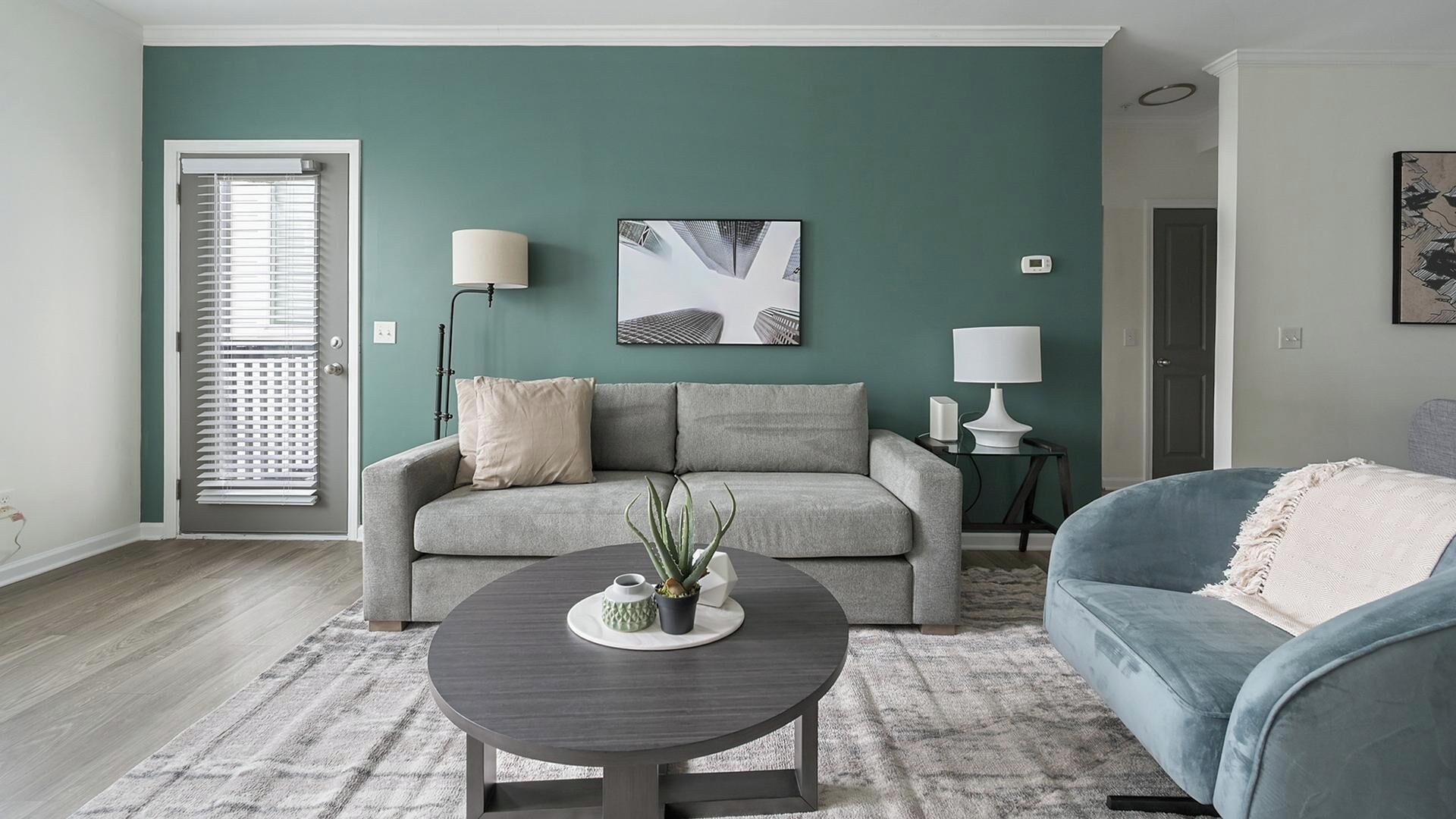 A living room with a grey sofa, a blue armchair, a round coffee table with a plant, and a painting on the wall.
