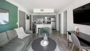 Uma sala moderna com um sofá cinzento, uma mesa de centro redonda e uma poltrona cinzenta. Uma cozinha com armários cinzentos e eletrodomésticos de aço inoxidável é visível ao fundo.