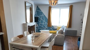 A modern dining area with a wooden table, white chairs, and a marble chair. A fireplace is visible in the background, and a sofa with cushions is placed near a window with curtains. - Large city apartment with 2 private indoor parking spaces (Beuvrages)